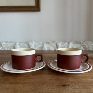 May include: Two matching coffee cups and saucers. The cups are a deep red with a cream-coloured rim. The saucers are white with a thin red border. The cups have a square handle. The set is on a wooden table.