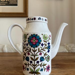 May include: White ceramic teapot with a floral pattern in blue, pink, and green. It features a curved handle and spout. The lid is adorned with small floral motifs.