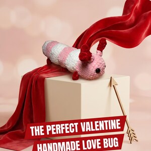 May include: A crocheted caterpillar plush with alternating pink and white segments, dark red antennae, and black eyes. A red velvet fabric drapes over a beige cube next to a gold arrow. Text on the image reads: THE PERFECT VALENTINE HANDMADE LOVE BUG.