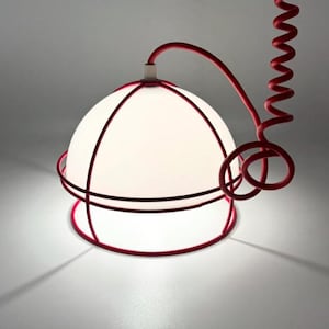 May include: A modern pendant light with a white, spherical shade encased in a red metal frame. The red cord, coiled in a spiral, adds a unique design element. The light casts a soft glow, creating a contemporary aesthetic.