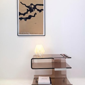 May include: A modern, clear acrylic bar cart with three shelves and wheels. The cart has a brown tint and features circular cutouts on the top shelf. A white lamp and a stack of books are on the cart. An abstract art print hangs on the wall.