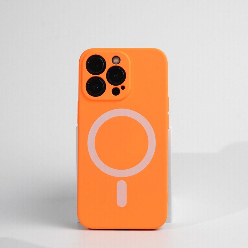 May include: Orange phone case with a white magnetic ring on the back. The case has a cutout for the camera lenses.