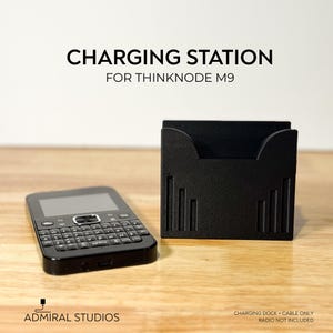 May include: A black charging station for Thinknode M9 with a rectangular base and vertical slot details, positioned next to a black handheld radio with a QWERTY keyboard and screen. Text reads CHARGING STATION FOR THINKNODE M9 and ADMIRAL STUDIOS.