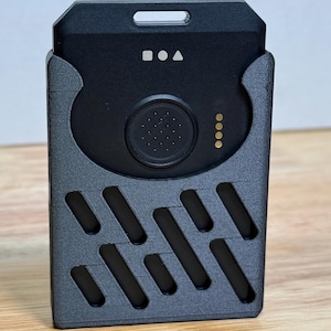 Peut inclure: Un appareil rectangulaire noir avec un bouton circulaire arrondi et quatre points dorés. L'appareil est logé dans un support gris à motifs avec une série de découpes diagonales. Le haut comporte un petit trou rectangulaire et trois petits symboles.