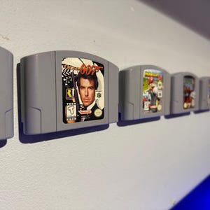 May include: A row of eight grey Nintendo 64 game cartridges mounted on a white wall. The cartridges feature various game titles, including "GoldenEye 007", "Mario Kart 64", and "Super Mario 64".