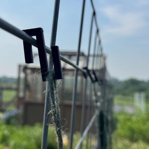 May include: Close-up of a metal garden fence with black clips and a length of twine. The fence is made of gray metal and is set against a backdrop of green foliage and a blue sky. The twine is knotted.