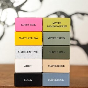 Puede incluir: Una pila de diez bloques rectangulares de varios colores, cada uno etiquetado con su nombre de color: Lotus Pink, Matte Yellow, Marble White, White, Black, Matte Bamboo Green, Matte Green, Olive Green, Matte Beige y Matte Blue.