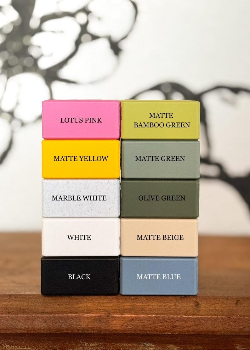 Puede incluir: Una pila de diez bloques rectangulares de varios colores, cada uno etiquetado con su nombre de color: Lotus Pink, Matte Yellow, Marble White, White, Black, Matte Bamboo Green, Matte Green, Olive Green, Matte Beige y Matte Blue.