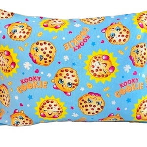 May include: A blue pillow with a repeating pattern of smiling cookie characters. The cookies are brown with chocolate chips and have pink cheeks and eyelashes. The background is light blue with yellow suns and white stars. The text "Kooky Cookie" is repeated on the pillow.