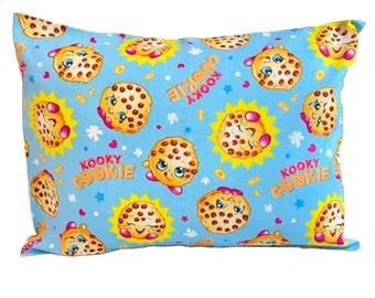 May include: A blue pillow with a repeating pattern of smiling cookie characters. The cookies are brown with chocolate chips and have pink cheeks and eyelashes. The background is light blue with yellow suns and white stars. The text "Kooky Cookie" is repeated on the pillow.