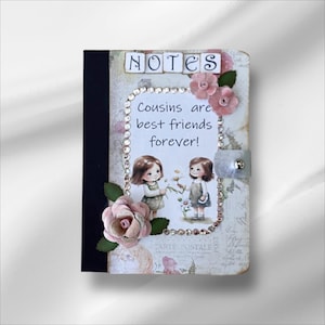 May include: A decorative notebook with the word "NOTES" at the top. The notebook features a design with two cartoon girls, pink flowers, and the text "Cousins are best friends forever!" The notebook has a black spine and a silver clasp.