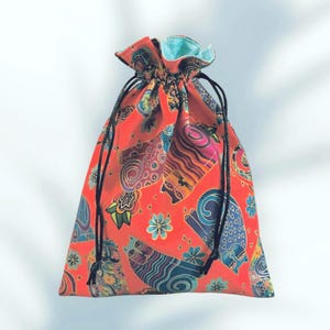 May include: A drawstring pouch featuring a vibrant orange background with a repeating pattern of stylized cats in shades of blue, purple, and gold, accented by small floral motifs. The bag has a light blue interior lining and black cord ties.