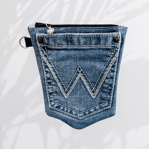 May include: A denim pouch with a zipper closure and a heart-shaped zipper pull. The pouch is made from a repurposed jean pocket, featuring white stitching and a signature "W" design. A black strap with a silver ring is attached.