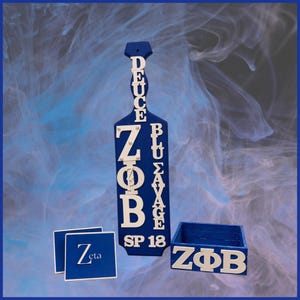 May include: A blue wooden paddle featuring white lettering that reads DEUCE, ZΦB, BLU SAVAGE, and SP 18. Beside it are two blue square coasters with the word Zeta in white, and a blue wooden box with white ZΦB lettering on the front side.