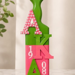 Peut inclure: Une pagaie en bois verticale peinte en vert et rose, ornée des lettres grecques Alpha, Kappa et Alpha. La lettre supérieure est décorée de perles blanches. La partie inférieure affiche les chiffres 19 et 08 peints en blanc sur fond rose.