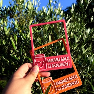 Könnte beinhalten: Zwei rechteckige Rahmen aus Kunststoff, einer in leuchtendem Pink und einer in Orange. Jeder Rahmen zeigt ein erhabenes Motiv eines Glases mit einem Herz auf der Vorderseite und den Schriftzug MARMELADEN GLASMOMENT in weißen Buchstaben unten.