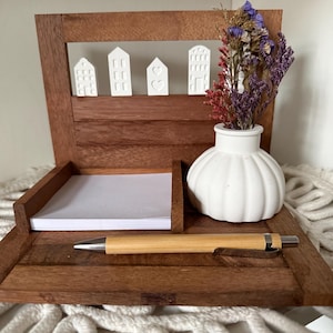 Könnte beinhalten: Ein Schreibtisch-Organizer aus Holz mit Notizblock, Stift und dekorativen Keramikhäusern. Eine kleine weiße Vase enthält Trockenblumen. Der Organizer ist aus dunklem Holz gefertigt und steht auf einer weißen Makramee-Oberfläche.