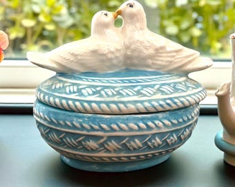 May include: A ceramic trinket box with a blue and white design. Two white doves are perched on the lid, their beaks touching. The box has a round shape with a detailed pattern around the sides.