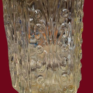 May include: Clear glass jar with a decorative pattern of swirls and circular accents. The jar has a rectangular shape with rounded edges and a slightly textured surface. The glass is transparent, allowing a view of the contents.