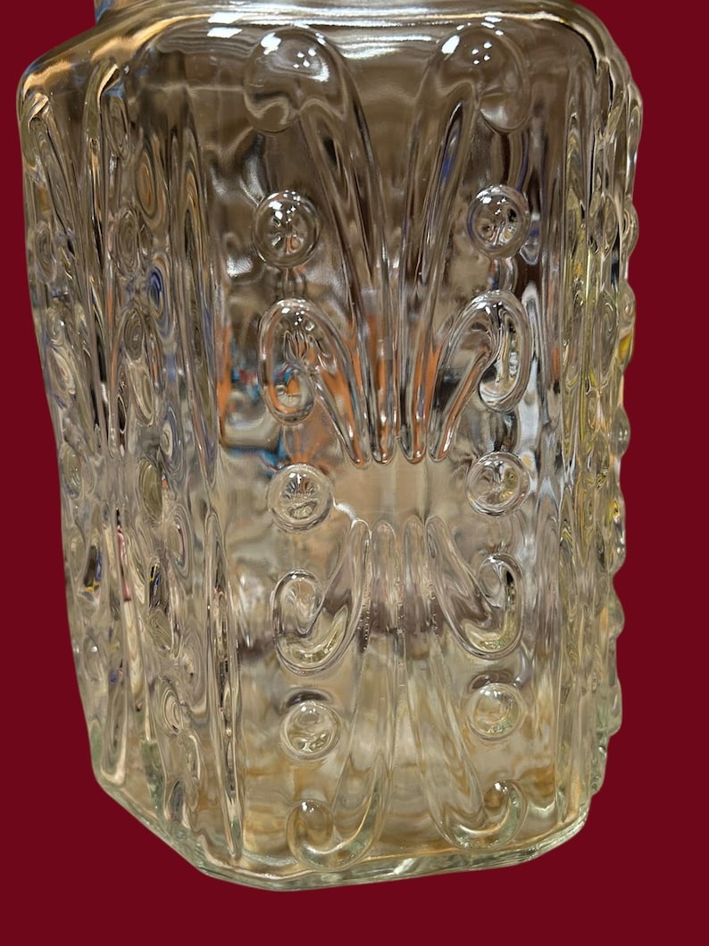 May include: Clear glass jar with a decorative pattern of swirls and circular accents. The jar has a rectangular shape with rounded edges and a slightly textured surface. The glass is transparent, allowing a view of the contents.