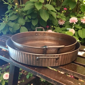 May include: Two antique copper pans, one nested inside the other, sit on a wooden bench. The larger pan has a ribbed edge and handles, while the smaller pan has a hammered texture. The background features green foliage and pink flowers.
