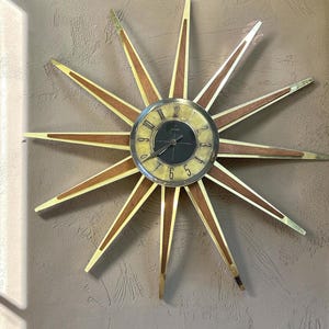 May include: A starburst wall clock featuring a central circular face with a black center and a light-colored dial with black numerals. The clock has twelve radiating spikes made of wood and brass-toned metal, extending outward from the center.