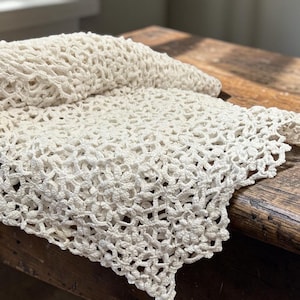 May include: An off-white, rectangular crochet runner featuring a repeating floral motif with an intricate, openwork lattice pattern. The textile has a scalloped edge and a soft, textured appearance, showcasing detailed needlework throughout the fabric.