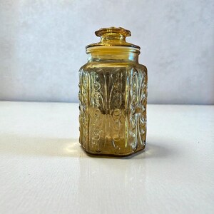 May include: Amber-colored glass jar with a lid. The jar has a rectangular shape with rounded corners and is decorated with a repeating floral pattern. The lid is also amber-colored and has a small knob on top. The jar is sitting on a white surface.