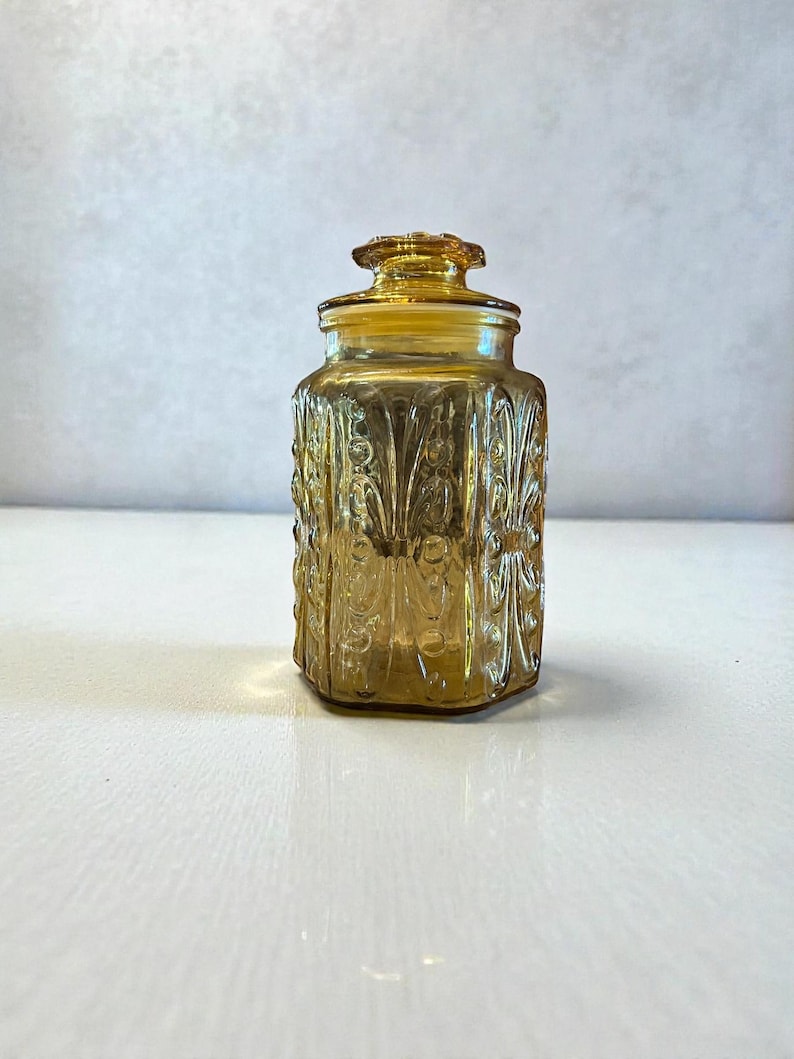 May include: Amber-colored glass jar with a lid. The jar has a rectangular shape with rounded corners and is decorated with a repeating floral pattern. The lid is also amber-colored and has a small knob on top. The jar is sitting on a white surface.