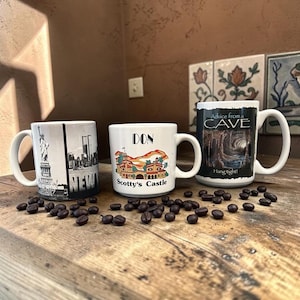 May include: Three ceramic mugs with handles. The left mug features a black and white Statue of Liberty and New York skyline. The center white mug displays a colorful illustration of Scotty's Castle with the text DON and Scotty's Castle. The right mug shows a cave interior with the text Advice from a CAVE Hang tight!