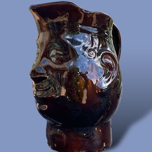 May include: A brown ceramic pitcher with a face sculpted on the front. The pitcher has a handle and a spout. The face has closed eyes, a nose, and a smiling mouth. The pitcher is glazed and has a glossy finish.