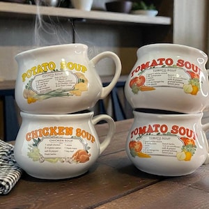 May include: Four white ceramic soup mugs stacked on a wooden table. Two mugs are labeled "Tomato Soup", one with a recipe graphic. The other two are labeled "Potato Soup" and "Chicken Soup", each with a recipe graphic.