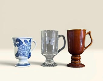 May include: Three decorative mugs. One is white with blue floral designs, another is clear glass with a butterfly, and the third is brown with a pedestal base. Each mug has a handle.