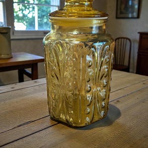 May include: A vintage amber glass jar with a lid. The jar has a decorative, embossed floral pattern and a hexagonal shape. The lid is also amber-colored and has a decorative design. The jar is sitting on a wooden table.