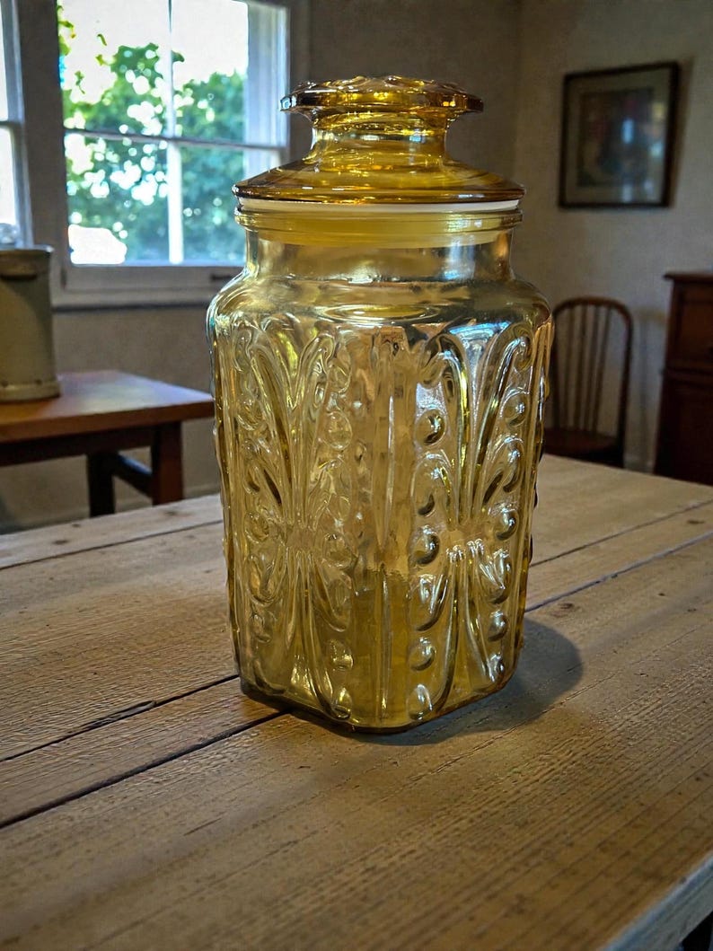 May include: A vintage amber glass jar with a lid. The jar has a decorative, embossed floral pattern and a hexagonal shape. The lid is also amber-colored and has a decorative design. The jar is sitting on a wooden table.