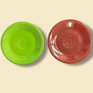 May include: Two ceramic plates, one in a vibrant green and the other in a rich red-brown hue. Both plates have a circular design with concentric rings and a central indentation. These plates are suitable for serving food.