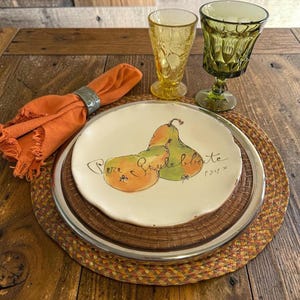 May include: A cream-colored ceramic plate with a hand-painted pear illustration and the text 'Pere Gulebbate P347', accompanied by an orange cloth napkin, a yellow glass tumbler, and a green textured glass goblet on a woven mat.