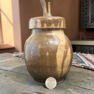May include: A round, brown ceramic jar with a matching lid featuring a tall, knob-like handle. The glaze has a mottled, dripped appearance with lighter tan streaks. A silver-colored United States quarter coin is placed at the base for scale.