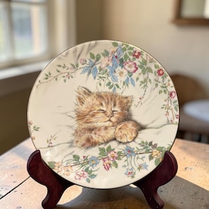 May include: A round ceramic decorative plate featuring a central illustration of a sleeping ginger tabby kitten. The kitten is surrounded by a delicate border of colorful flowers, including pink, blue, and yellow blossoms with green leaves.