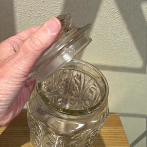 May include: A clear glass jar with a decorative floral pattern and a matching lid. The jar is open, revealing the interior. The jar is sitting on a wooden surface.