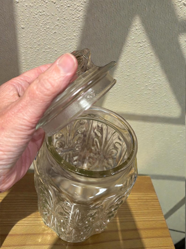 May include: A clear glass jar with a decorative floral pattern and a matching lid. The jar is open, revealing the interior. The jar is sitting on a wooden surface.