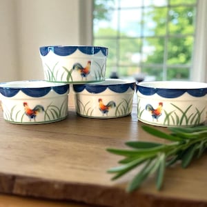 May include: Four white ceramic ramekins with a blue scalloped rim and a rooster design. The ramekins have a rooster illustration, green grass, and a blue scalloped border. They are arranged on a wooden surface.