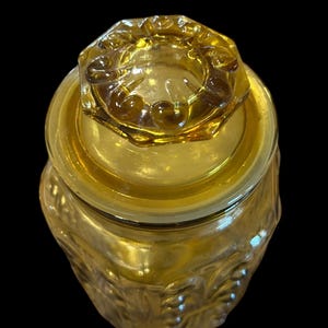 May include: Amber-colored glass jar with a lid. The jar has a textured, embossed design and a matching lid with a decorative rim. The jar is likely for storage or display.