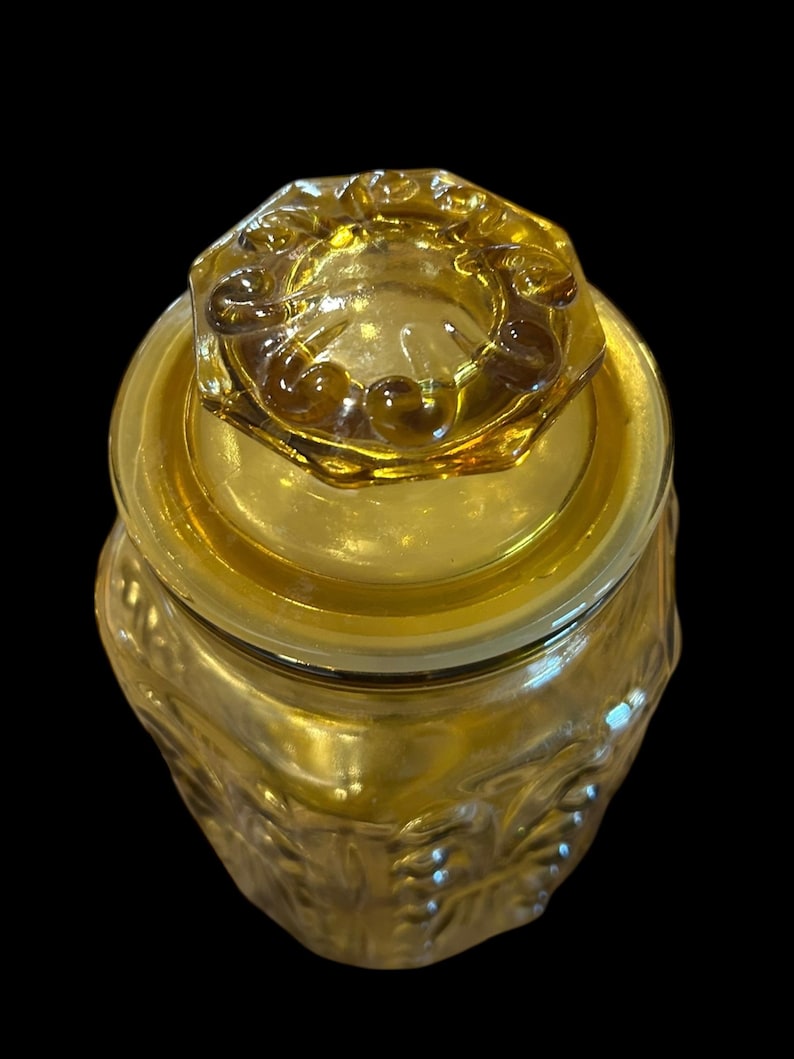 May include: Amber-colored glass jar with a lid. The jar has a textured, embossed design and a matching lid with a decorative rim. The jar is likely for storage or display.