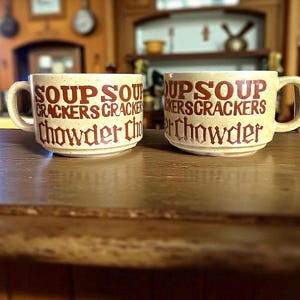 May include: Two cream-colored soup mugs with brown speckled text that reads "SOUP CRACKERS Chowder". The mugs have large handles and are sitting on a wooden surface. The background is a blurred wooden interior.