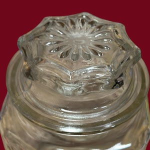 May include: Clear glass jar with a decorative, star-shaped lid. The lid features a detailed, embossed floral design. The jar's body has a textured surface, and the glass appears to be thick and sturdy. The background is a solid, dark red.