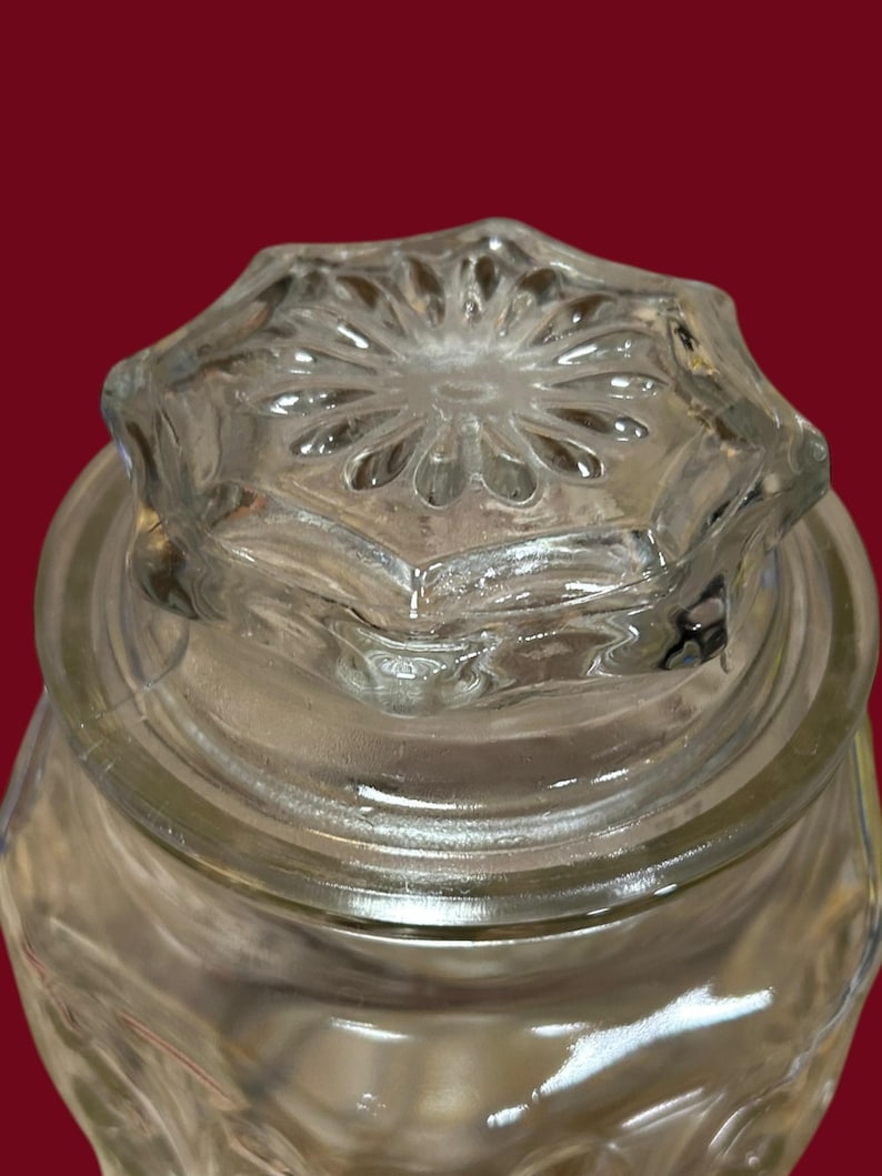 May include: Clear glass jar with a decorative, star-shaped lid. The lid features a detailed, embossed floral design. The jar's body has a textured surface, and the glass appears to be thick and sturdy. The background is a solid, dark red.