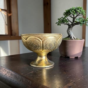 May include: A gold-colored, ornate bowl with a pedestal base. The bowl features detailed embossed patterns and sits on a dark wooden surface. A small bonsai tree in a brown pot is to the right of the bowl.