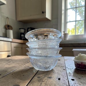 May include: A stack of four clear glass bowls with embossed floral patterns. The bowls are stacked on a rustic wooden table. The bowls are in a kitchen setting with a window in the background.