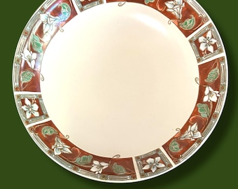 May include: A round, cream-colored plate with a decorative border. The border features a repeating pattern of white flowers, green leaves, and brown rectangular panels. The plate has a wide, empty center.